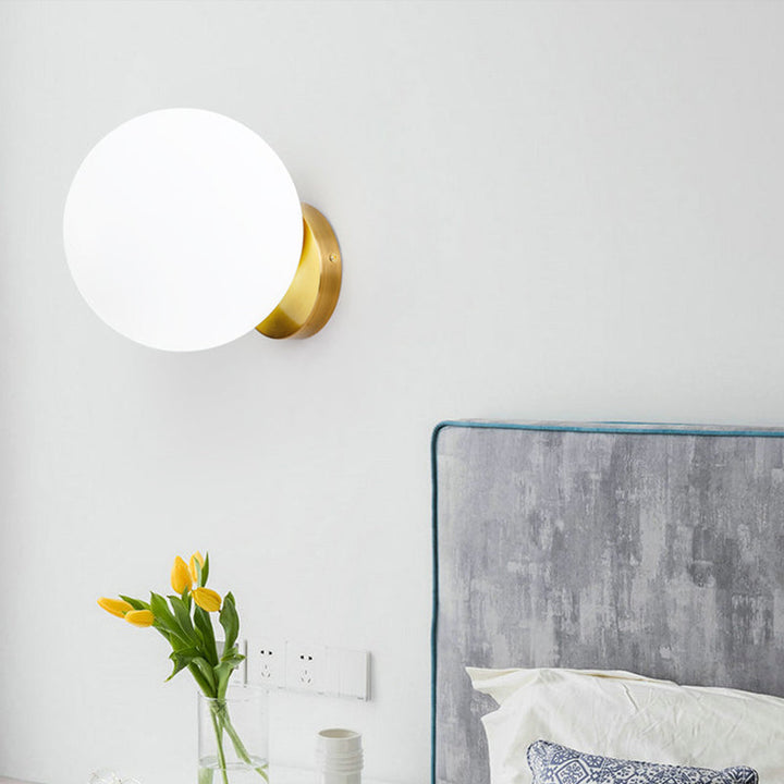 Wandlampe mit milchiger Glaskugel über Bett mit grauem Kopfteil und frischen Blumen

