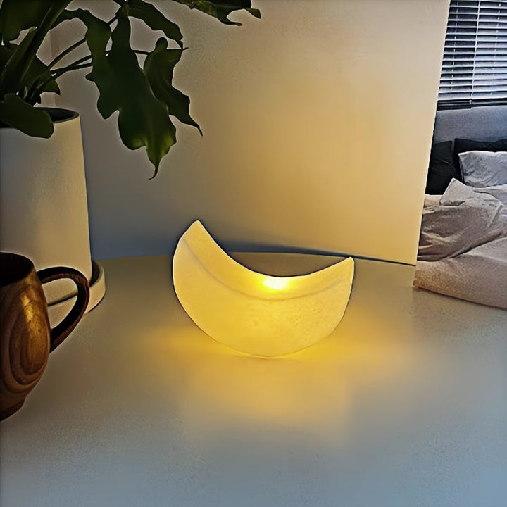 Leuchtende Mondlampe in sanftem Gelb auf weißer Kommode mit Pflanzen im Hintergrund

